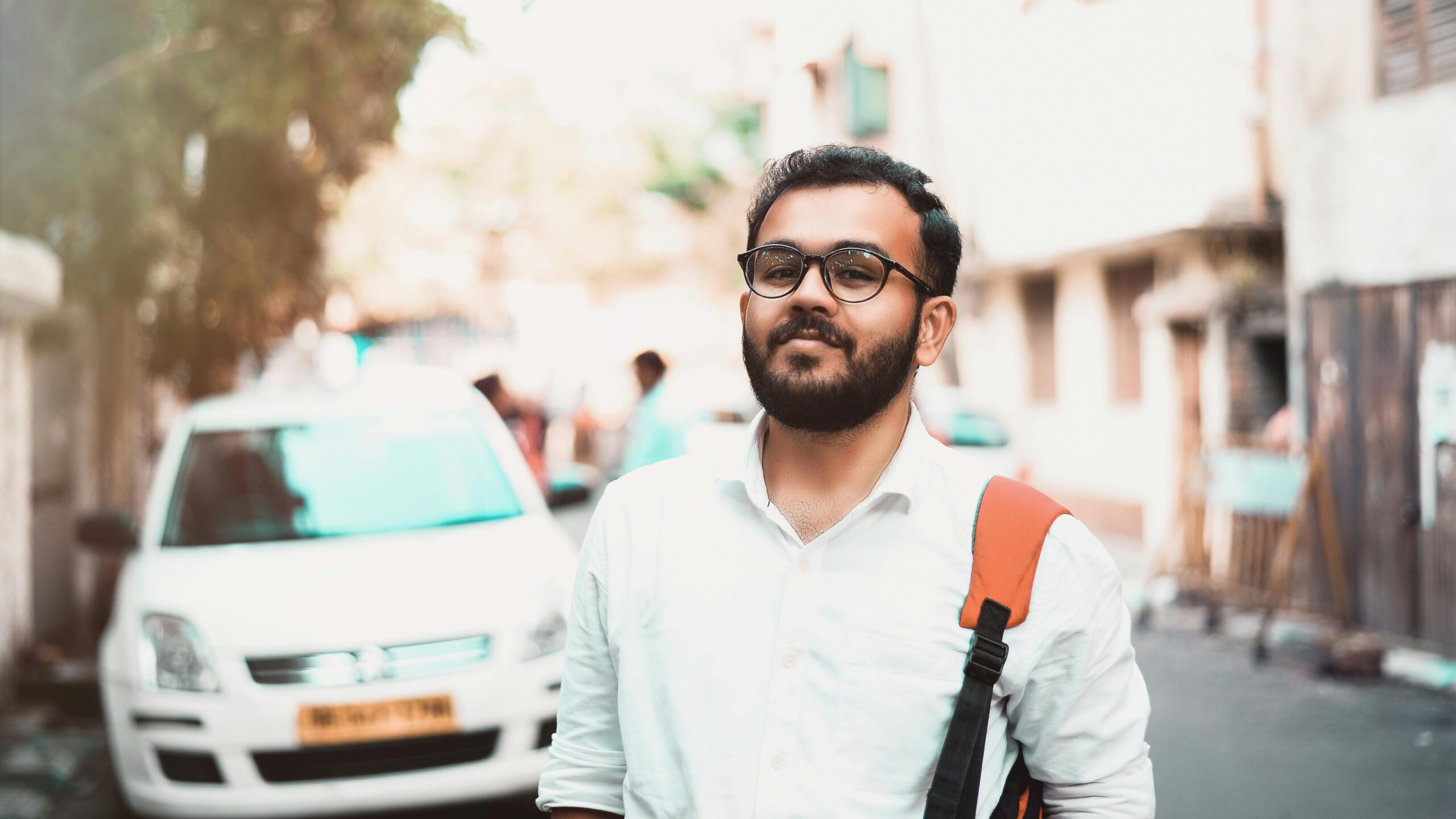 Confident man with glasses and backpack on a sunny urban street with cars in the background.